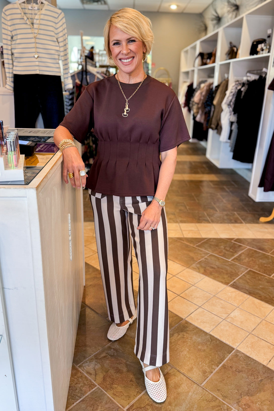 A woman stands in a boutique wearing a dark brown short-sleeve peplum top and brown-and-white vertically striped wide-leg pants, paired with white mule flats.
