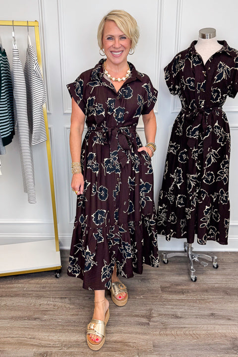 Front view of woman wearing Swagger Boutique’s Brown. &amp; Black Floral Print Midi Dress by Entro with clothing hanging in the background.
