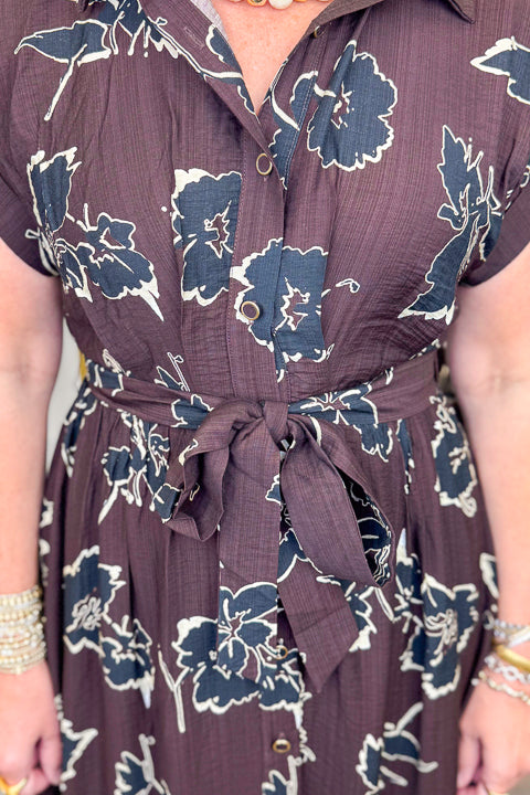 Front detail view of woman wearing Swagger Boutique’s Brown. &amp; Black Floral Print Midi Dress by Entro with clothing hanging in the background.