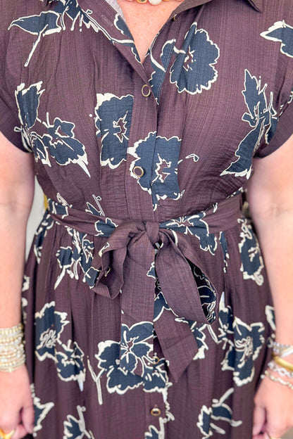 Front detail view of woman wearing Swagger Boutique’s Brown. &amp; Black Floral Print Midi Dress by Entro with clothing hanging in the background.