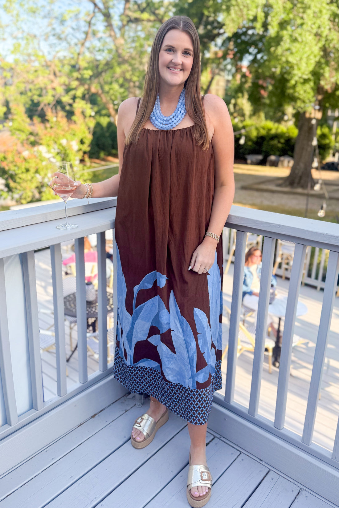 Front view of woman wearing Brown &amp; Blue Floral Maxi Dress with gold sandals outside at a restaurant patio.