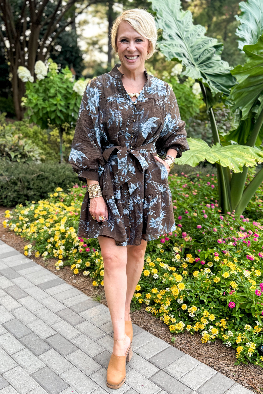 Front view of woman wearing Swagger Boutique’s Brown &amp; Blue Floral Tie Waist Mini Dress standing outside by a garden.