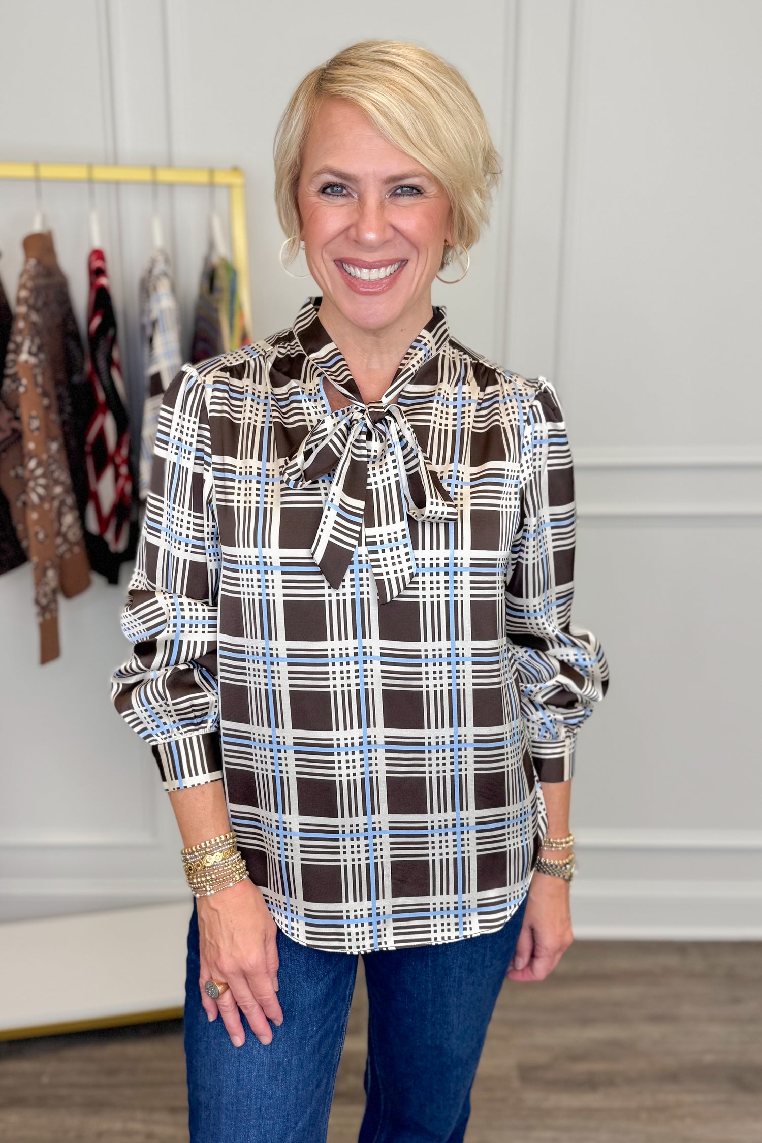 Front view of woman wearing Swagger Boutique’s Brown &amp; Blue Plaid Tie Neck Top with jeans with clothing hanging in the background.