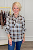 Front view of woman wearing Swagger Boutique’s Brown & Blue Plaid Tie Neck Top with jeans with clothing hanging in the background.