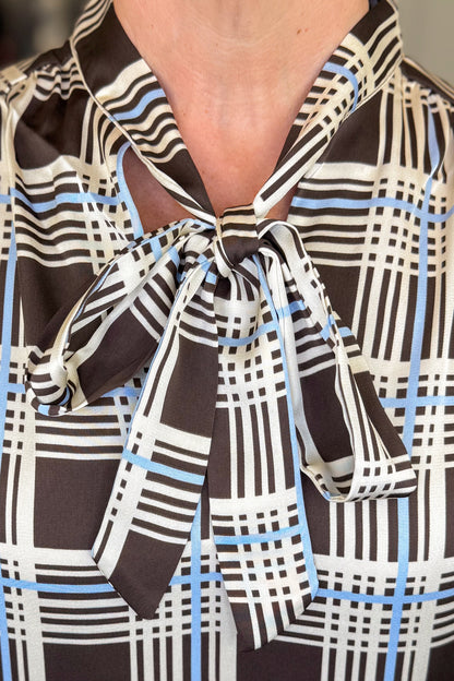 Front detail view of woman wearing Swagger Boutique’s Brown &amp; Blue Plaid Tie Neck Top with jeans with clothing hanging in the background.