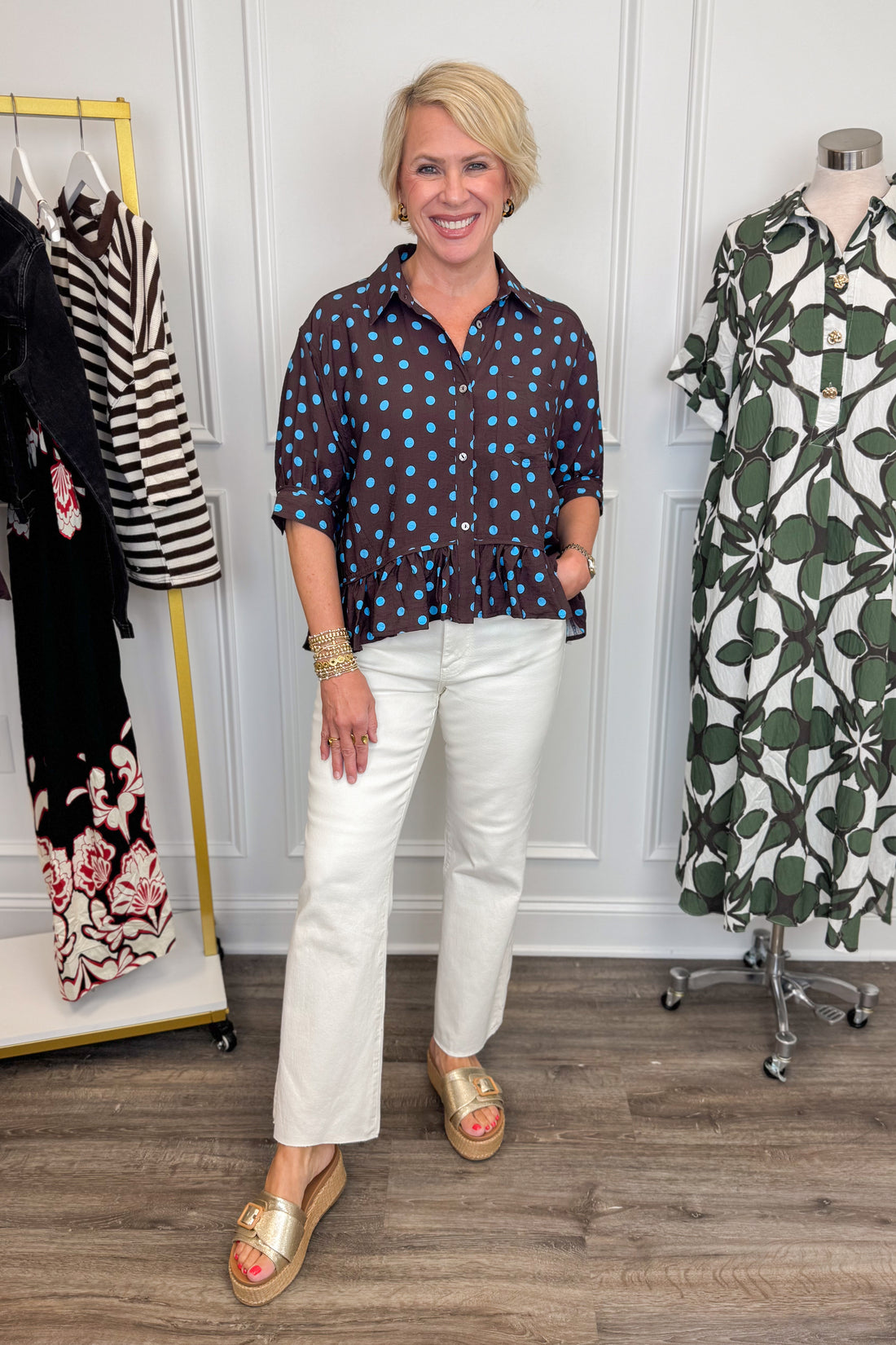 Front view of woman wearing Swagger Boutique’s Brown &amp; Blue Polka Dot Peplum Top by Entro with jeans with clothing hanging in the background. 