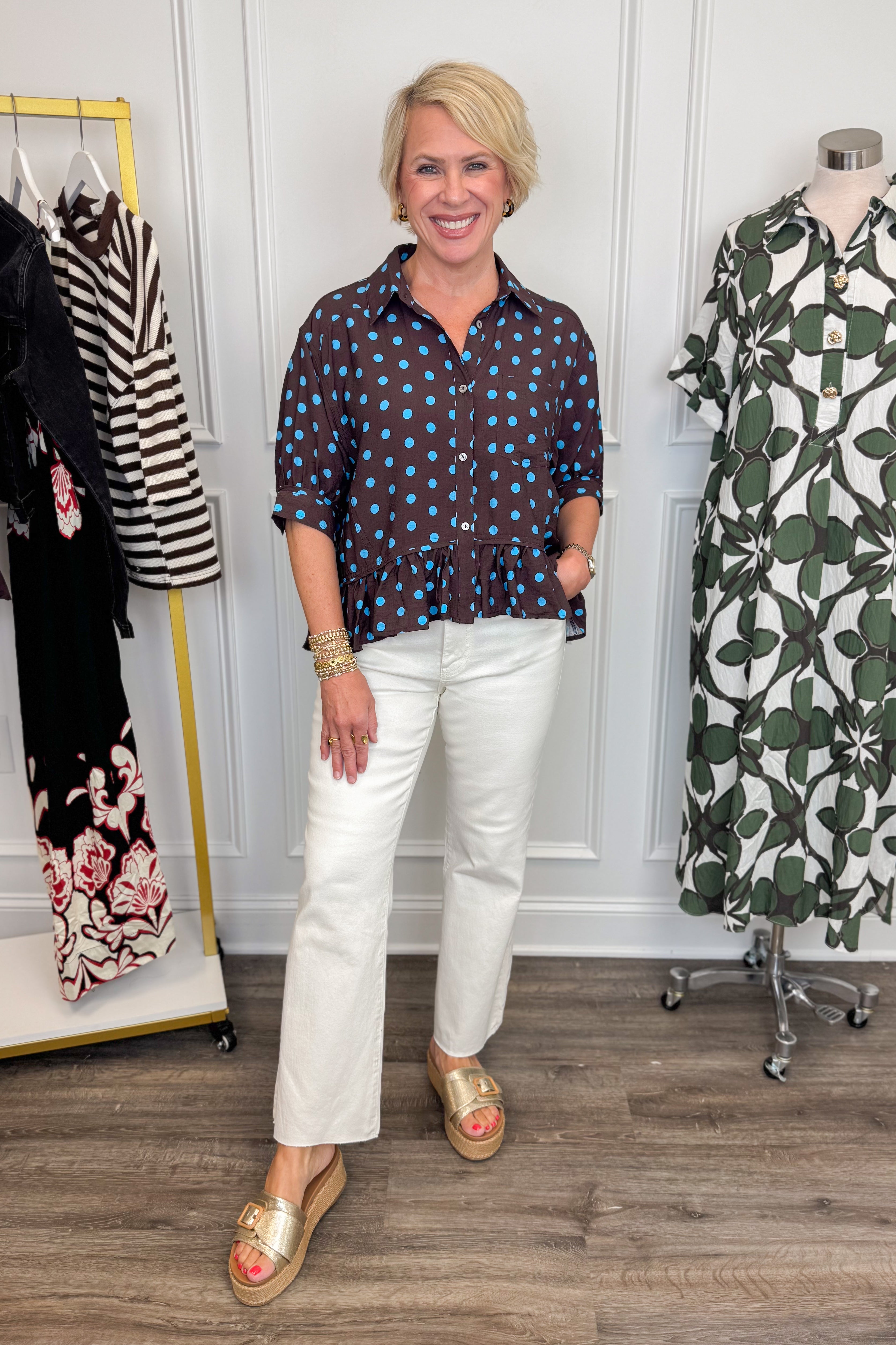 Front view of woman wearing Swagger Boutique’s Brown &amp; Blue Polka Dot Peplum Top by Entro with jeans with clothing hanging in the background. 