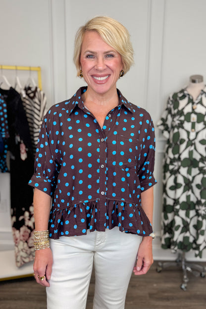 Front view of woman wearing Swagger Boutique’s Brown &amp; Blue Polka Dot Peplum Top by Entro with jeans with clothing hanging in the background. 