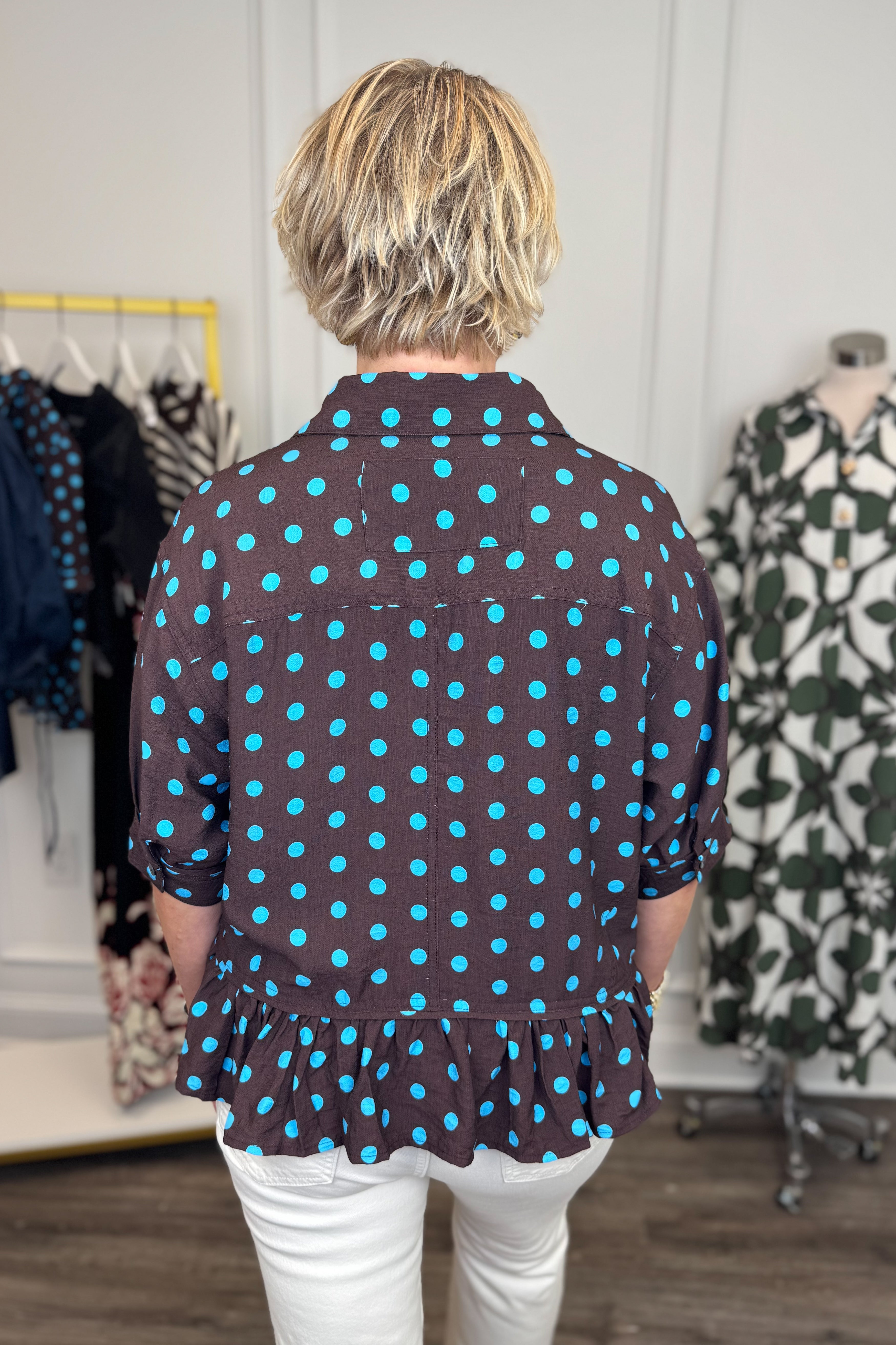 Back view of woman wearing Swagger Boutique’s Brown &amp; Blue Polka Dot Peplum Top by Entro with jeans with clothing hanging in the background. 