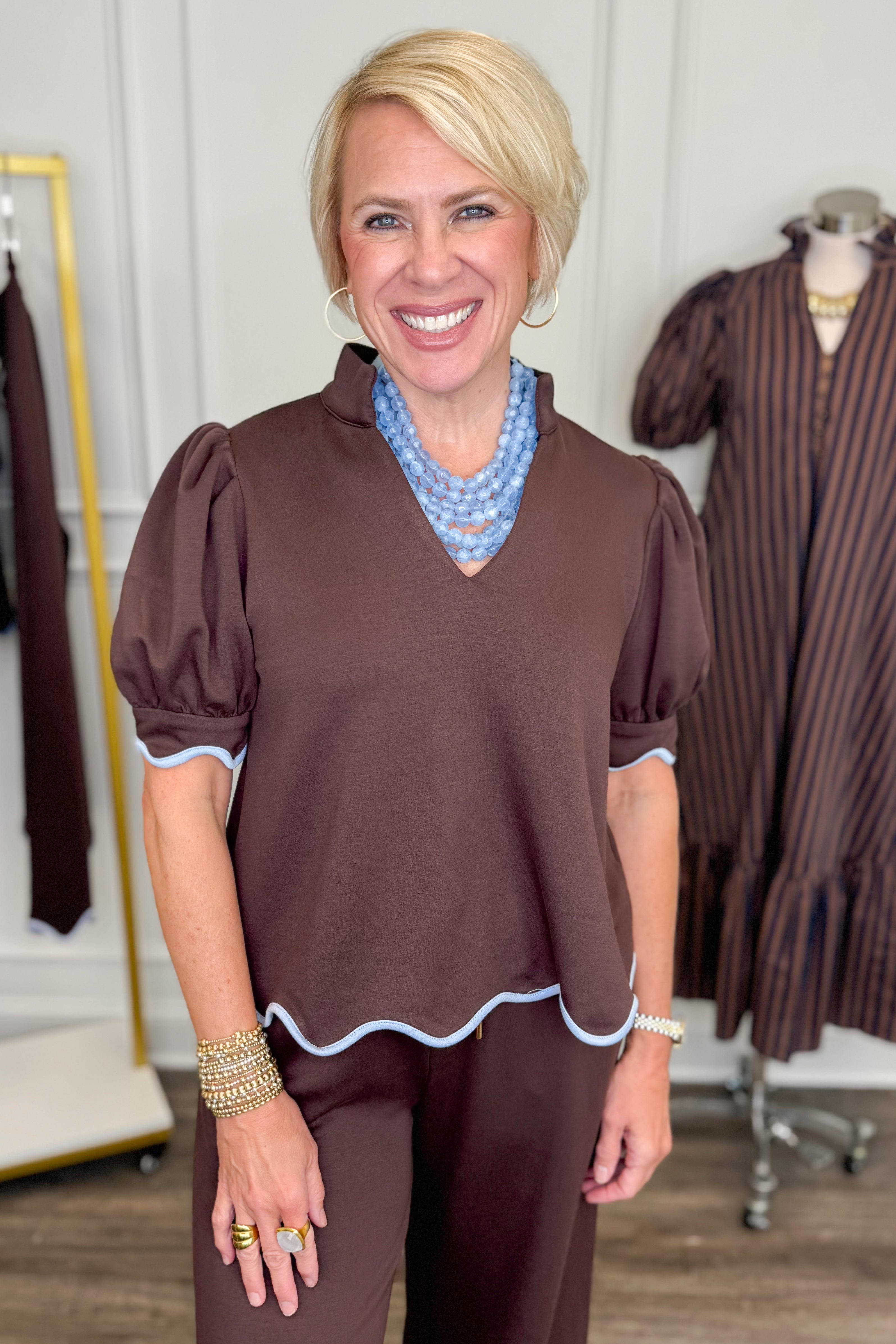 Front view of woman wearing Swagger Boutique’s Brown &amp; Blue Scalloped Top by Entro with matching pants with clothing hanging in the background.