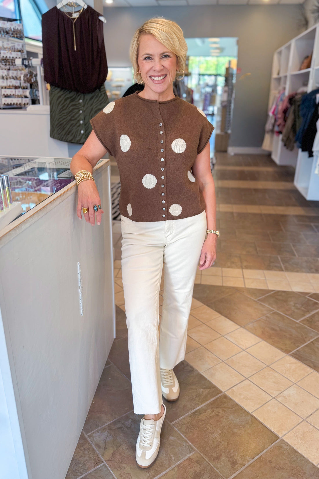 Front view of woman wearing Swagger Boutique’s Brown &amp; Cream Polka Dot Sleeveless Button Up Sweater by Entro with jeans standing at the store.