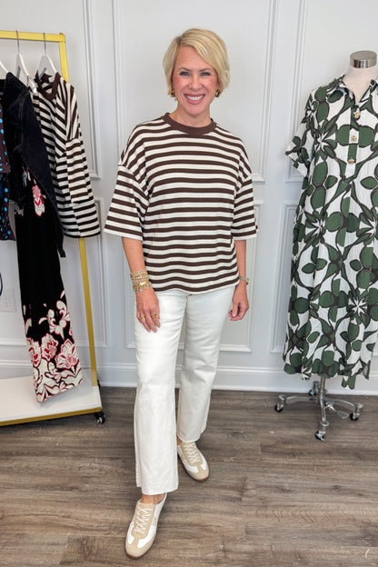 Front view of woman wearing Swagger Boutique’s Brown &amp; Cream Striped Textured Tee by Entro with jeans with clothing hanging in the background. 