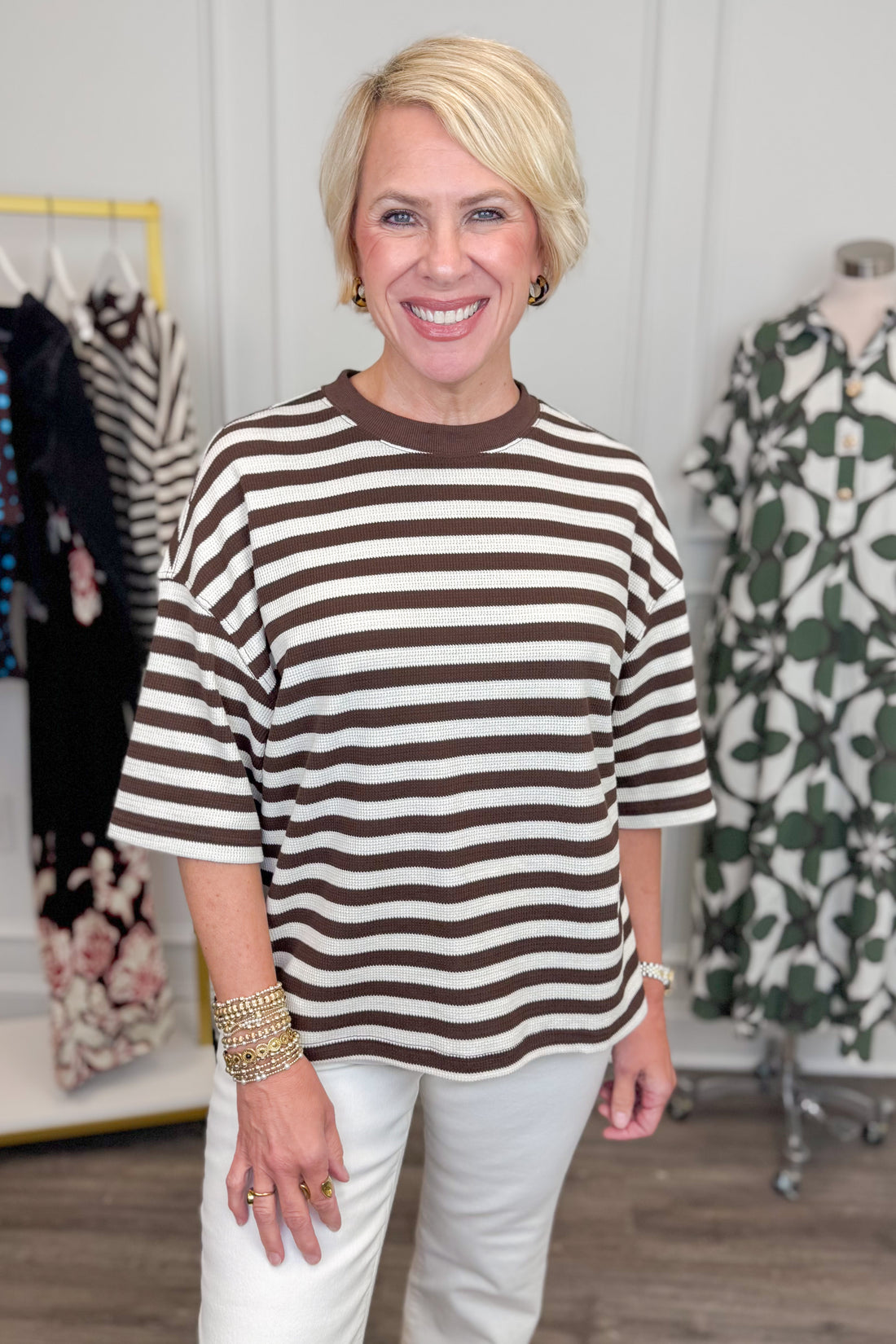 Front view of woman wearing Swagger Boutique’s Brown &amp; Cream Striped Textured Tee by Entro with jeans with clothing hanging in the background. 