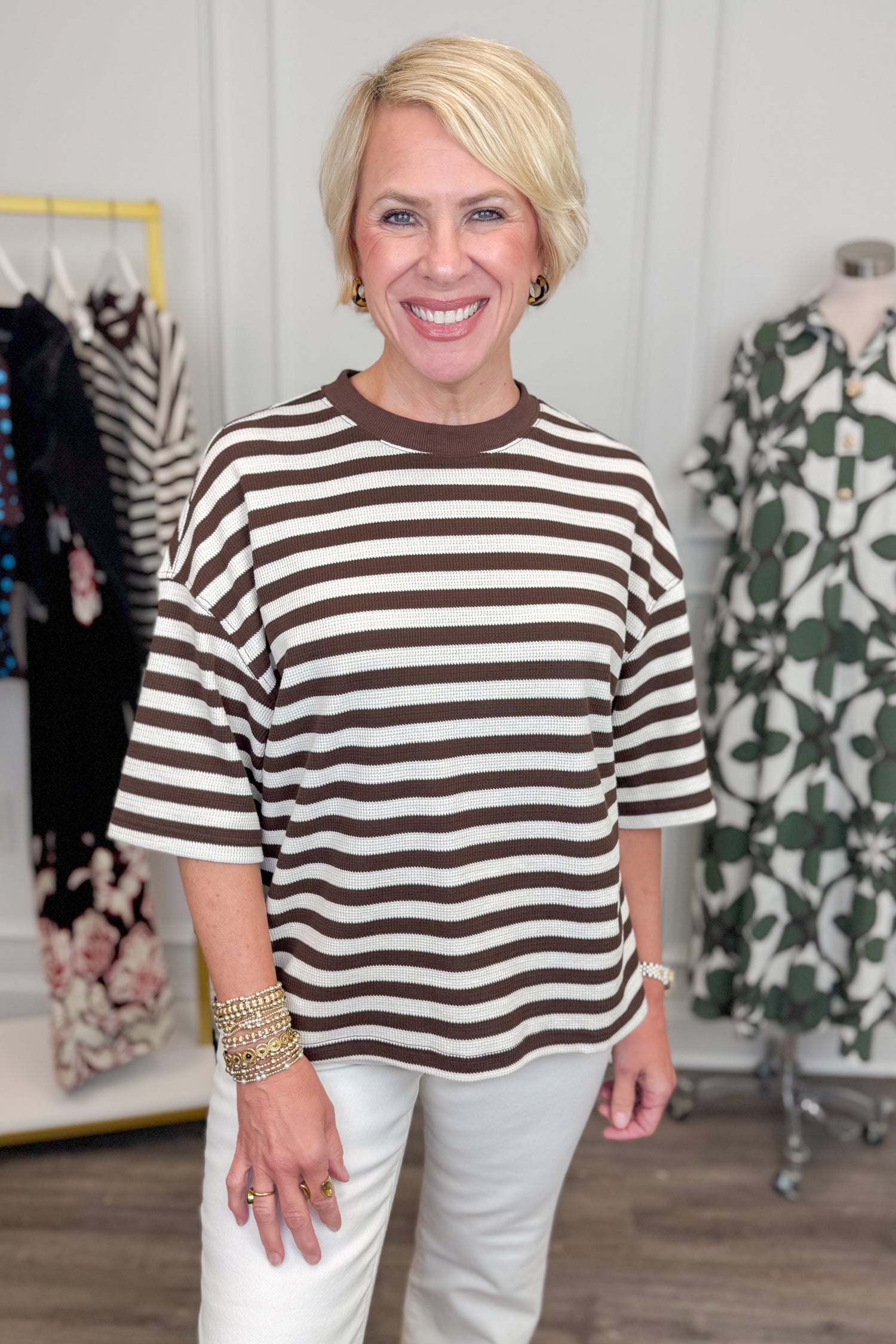 Front view of woman wearing Swagger Boutique’s Brown &amp; Cream Striped Textured Tee by Entro with jeans with clothing hanging in the background. 