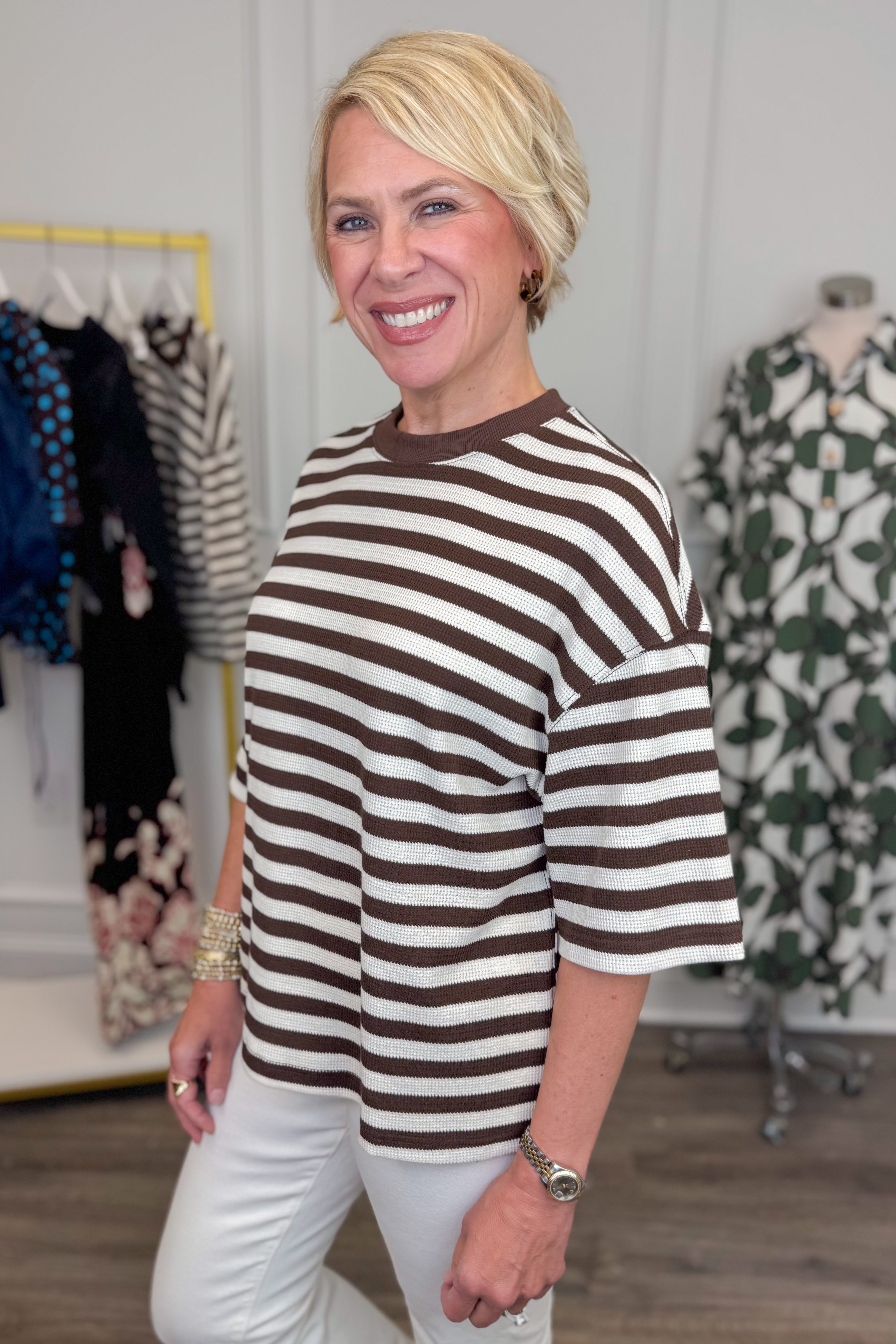 Side view of woman wearing Swagger Boutique’s Brown &amp; Cream Striped Textured Tee by Entro with jeans with clothing hanging in the background. 