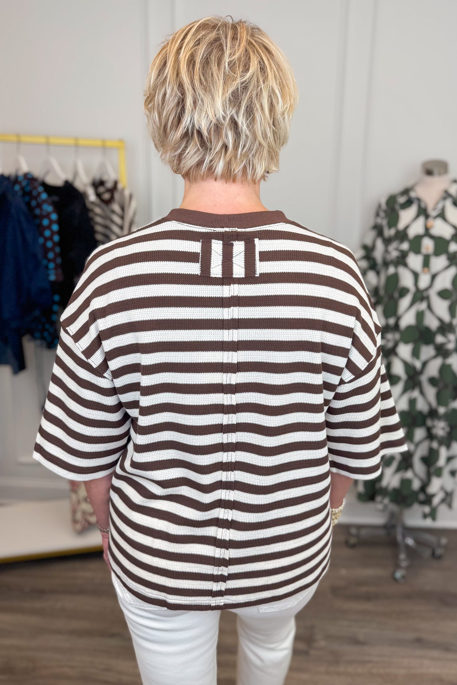 Back view of woman wearing Swagger Boutique’s Brown &amp; Cream Striped Textured Tee by Entro with jeans with clothing hanging in the background. 