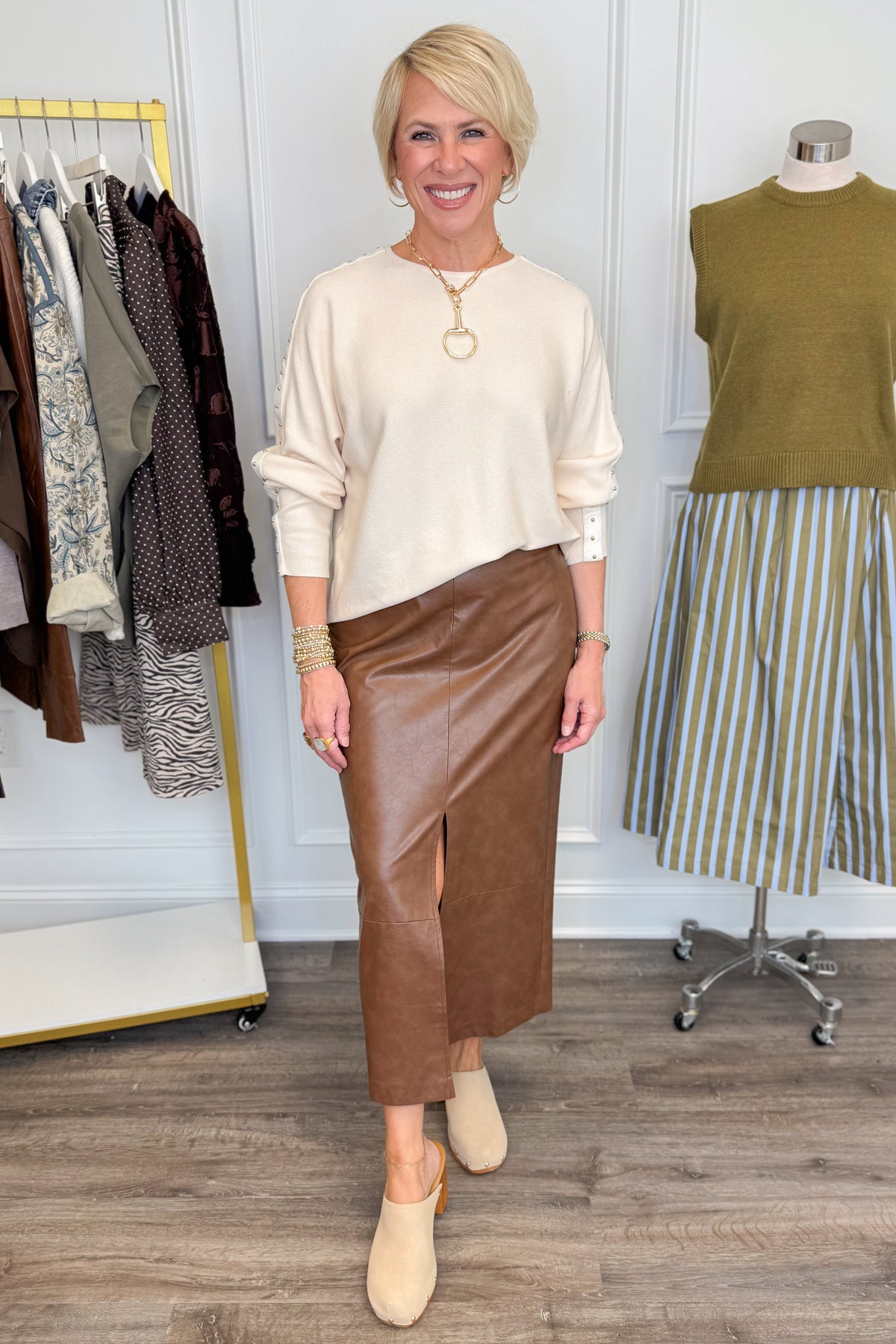 Front view of woman wearing Swagger Boutique’s Brown Faux Leather Slit Midi Skirt with Ecru Studded Sleeve Sweater with clothing hanging in the background.