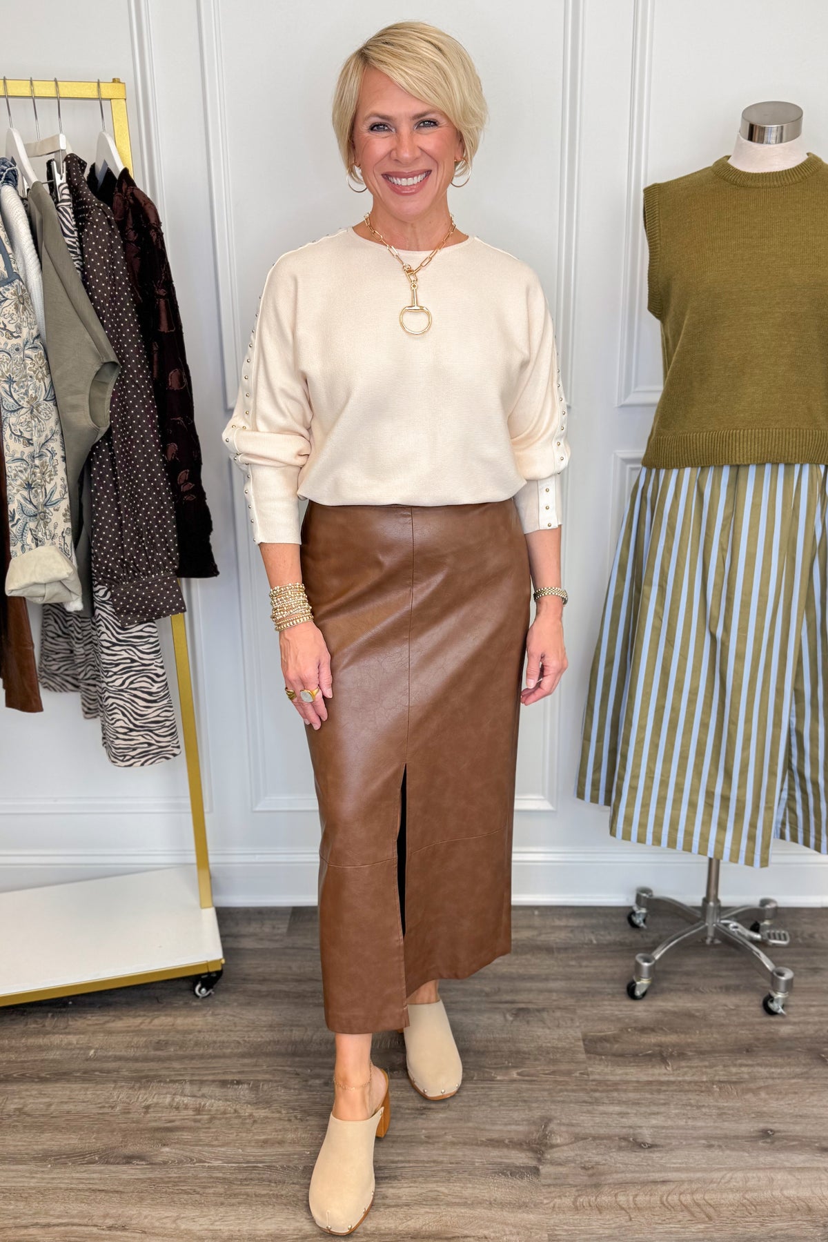 Front view of woman wearing Swagger Boutique’s Brown Faux Leather Slit Midi Skirt with Ecru Studded Sleeve Sweater with clothing hanging in the background.