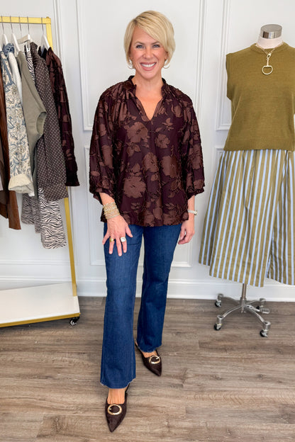 Front view of woman wearing Swagger Boutique’s Brown Floral &amp; Velvet 3/4 Sleeve Top with jeans with clothing hanging in the background.