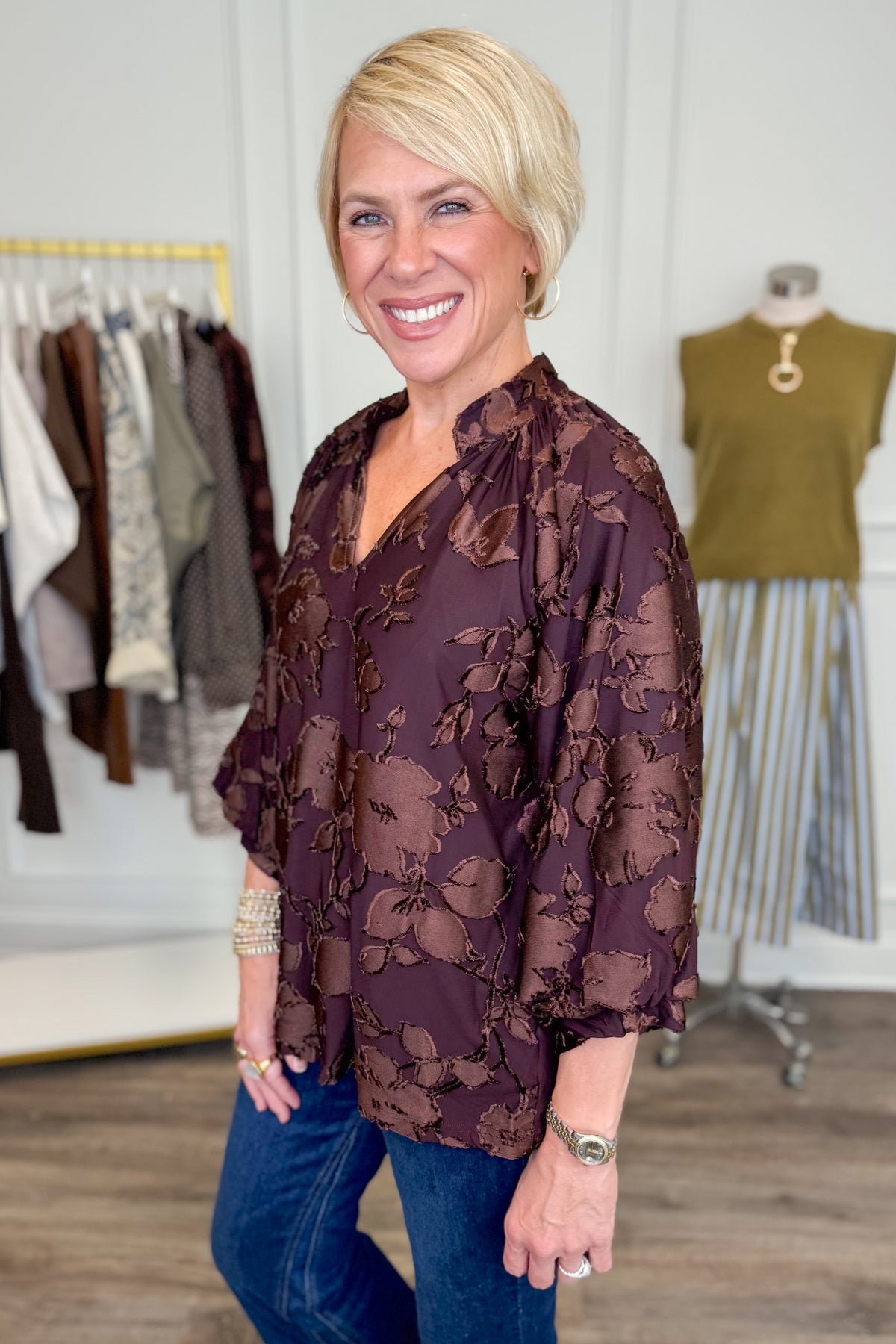 Side view of woman wearing Swagger Boutique’s Brown Floral &amp; Velvet 3/4 Sleeve Top with jeans with clothing hanging in the background.