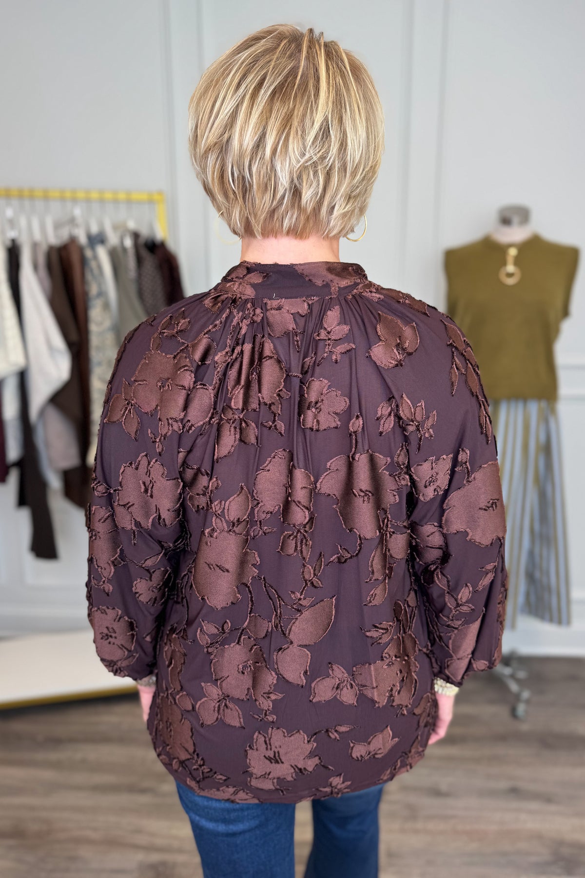 Back view of woman wearing Swagger Boutique’s Brown Floral &amp; Velvet 3/4 Sleeve Top with jeans with clothing hanging in the background.