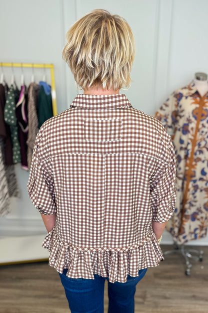 A woman viewed from behind wearing a brown and white gingham check shirt with a ruffled hem, paired with blue jeans, in a clothing boutique setting.