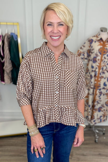 A smiling blonde woman stands indoors wearing a brown and white gingham button-front shirt with three-quarter sleeves and a peplum hem, paired with dark blue jeans. A clothing rack and a patterned dress on a mannequin are visible in the softly lit boutique setting.