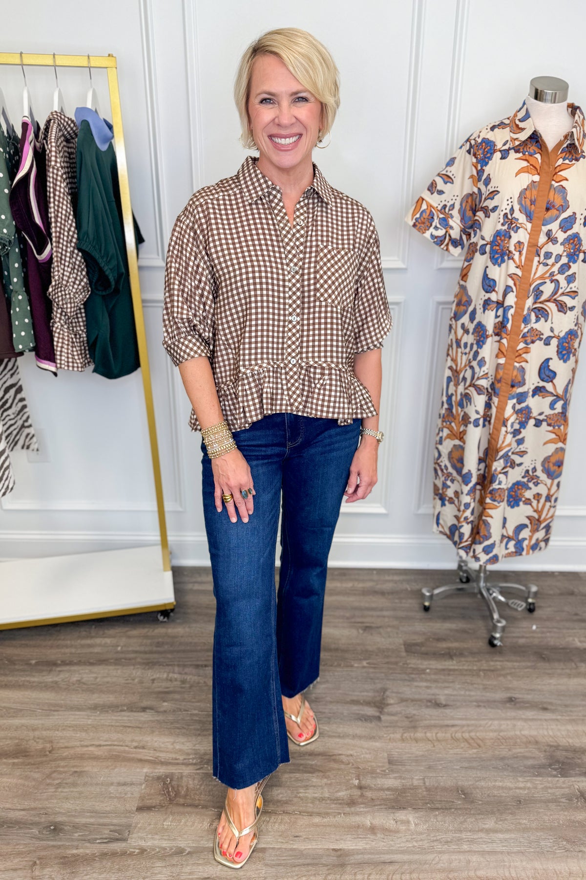 A woman stands in a boutique wearing a brown and beige gingham button-up shirt with dark blue jeans, while a patterned shirt is displayed on a mannequin in the background.