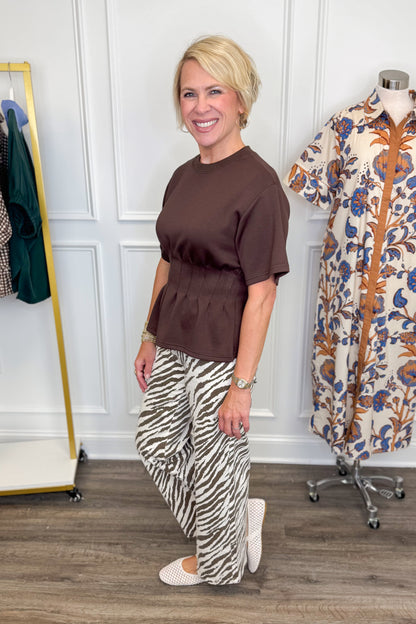 A woman stands in a boutique wearing a brown short-sleeve peplum top and beige-and-black zebra-print wide-leg pants, paired with white Mary Jane flats. A clothing rack and a mannequin wearing a floral shirt are visible in the background.