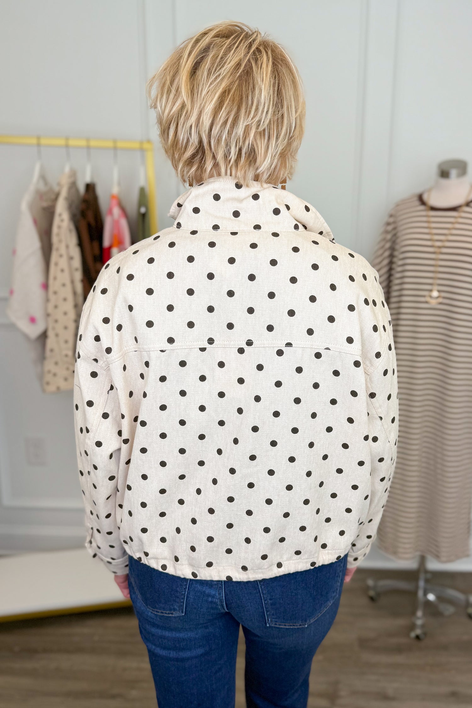 Back view of woman wearing Swagger Boutique’s Brown Polka Dot Denim Jacket with jeans with clothing hanging in the background.