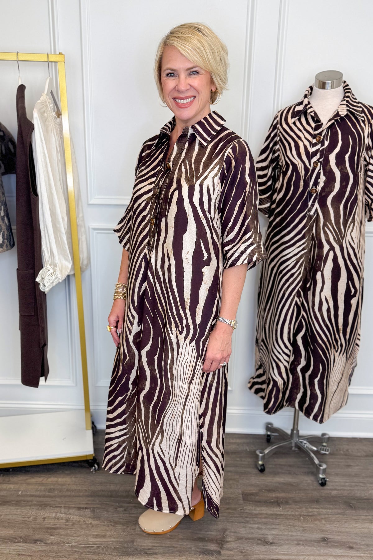 Side view of woman wearing Swagger Boutique’s Brown Zebra Midi Dress by Entro with clogs with clothing hanging in the background.