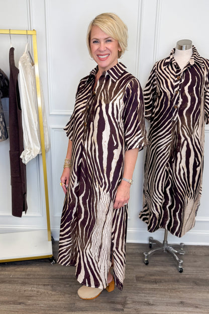 Side view of woman wearing Swagger Boutique’s Brown Zebra Midi Dress by Entro with clogs with clothing hanging in the background.