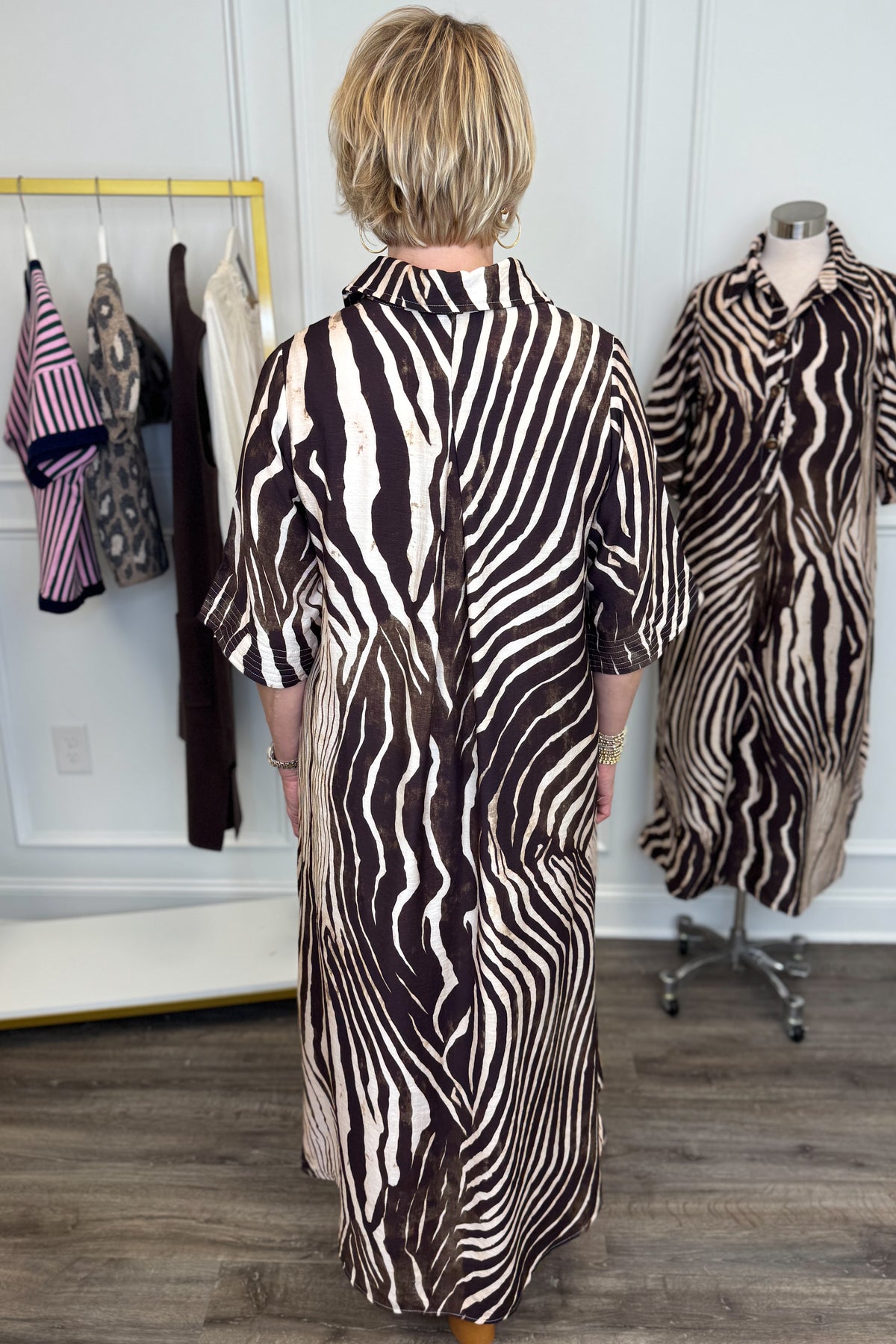 Back view of woman wearing Swagger Boutique’s Brown Zebra Midi Dress by Entro with clogs with clothing hanging in the background.