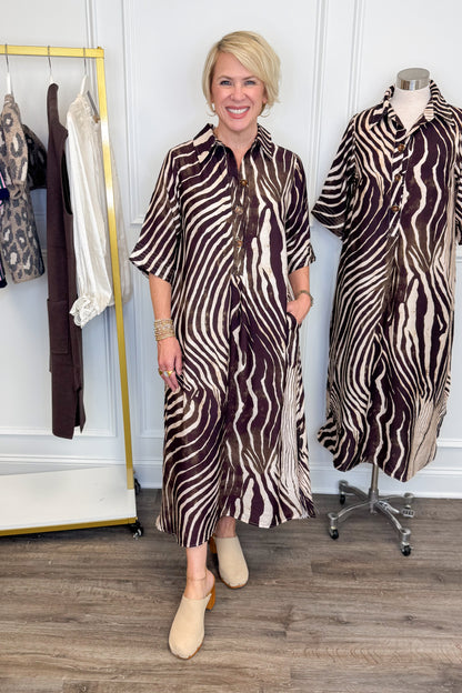 Front view of woman wearing Swagger Boutique’s Brown Zebra Midi Dress by Entro with clogs with clothing hanging in the background.