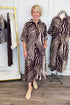 Front view of woman wearing Swagger Boutique’s Brown Zebra Midi Dress by Entro with clogs with clothing hanging in the background.