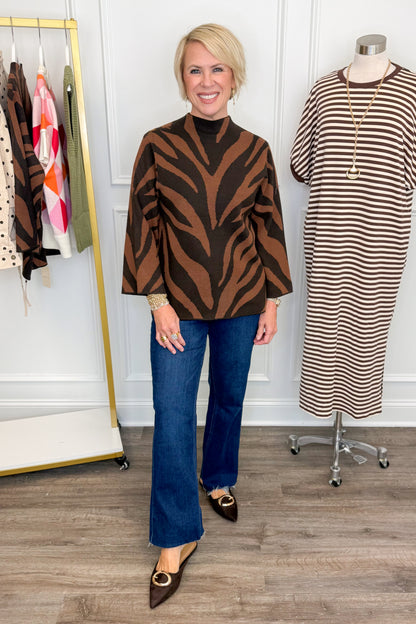 Front view of woman wearing Swagger Boutique’s Brown Zebra Mock Neck Sweater with jeans with clothing hanging in the background.