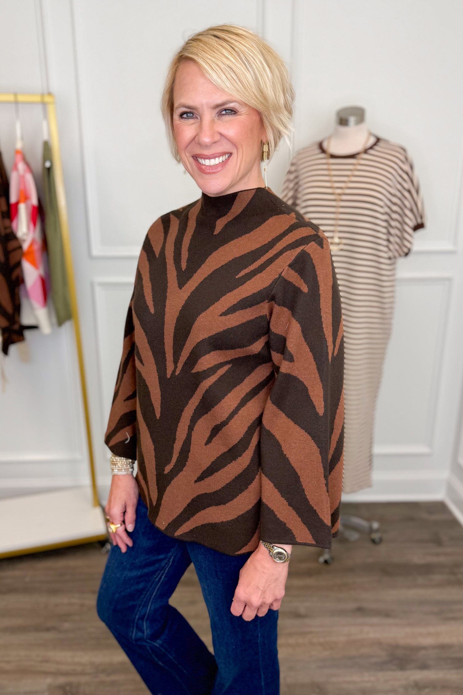 Side view of woman wearing Swagger Boutique’s Brown Zebra Mock Neck Sweater with jeans with clothing hanging in the background.