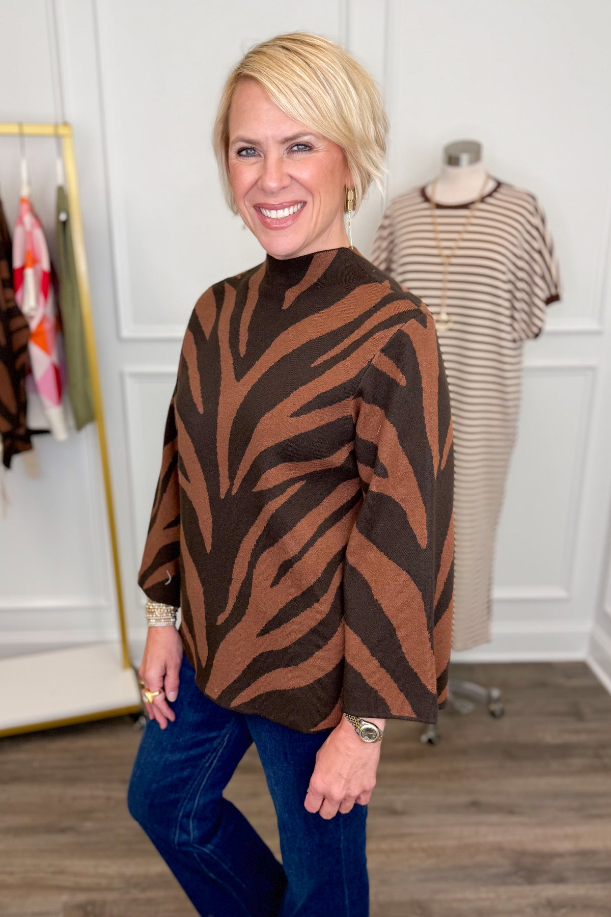 Side view of woman wearing Swagger Boutique’s Brown Zebra Mock Neck Sweater with jeans with clothing hanging in the background.
