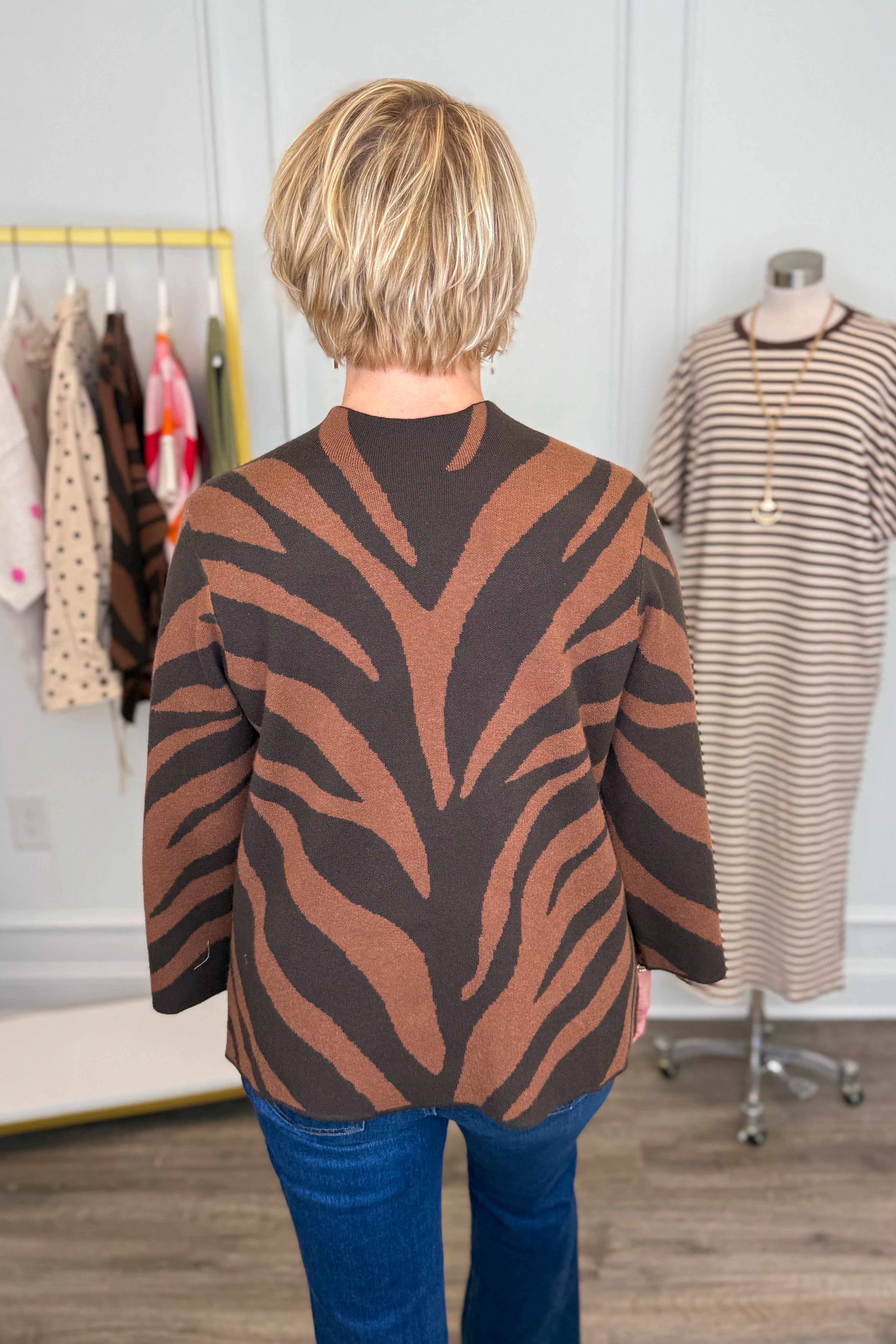 Back view of woman wearing Swagger Boutique’s Brown Zebra Mock Neck Sweater with jeans with clothing hanging in the background.