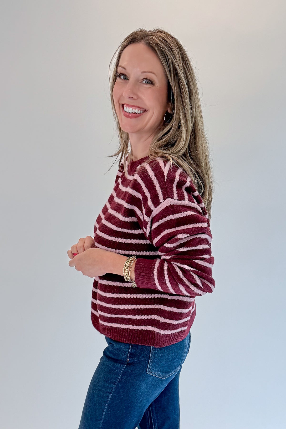 Side view of woman wearing Swagger Boutique’s Burgundy &amp; Pink Striped Sweater with jeans against white studio background.