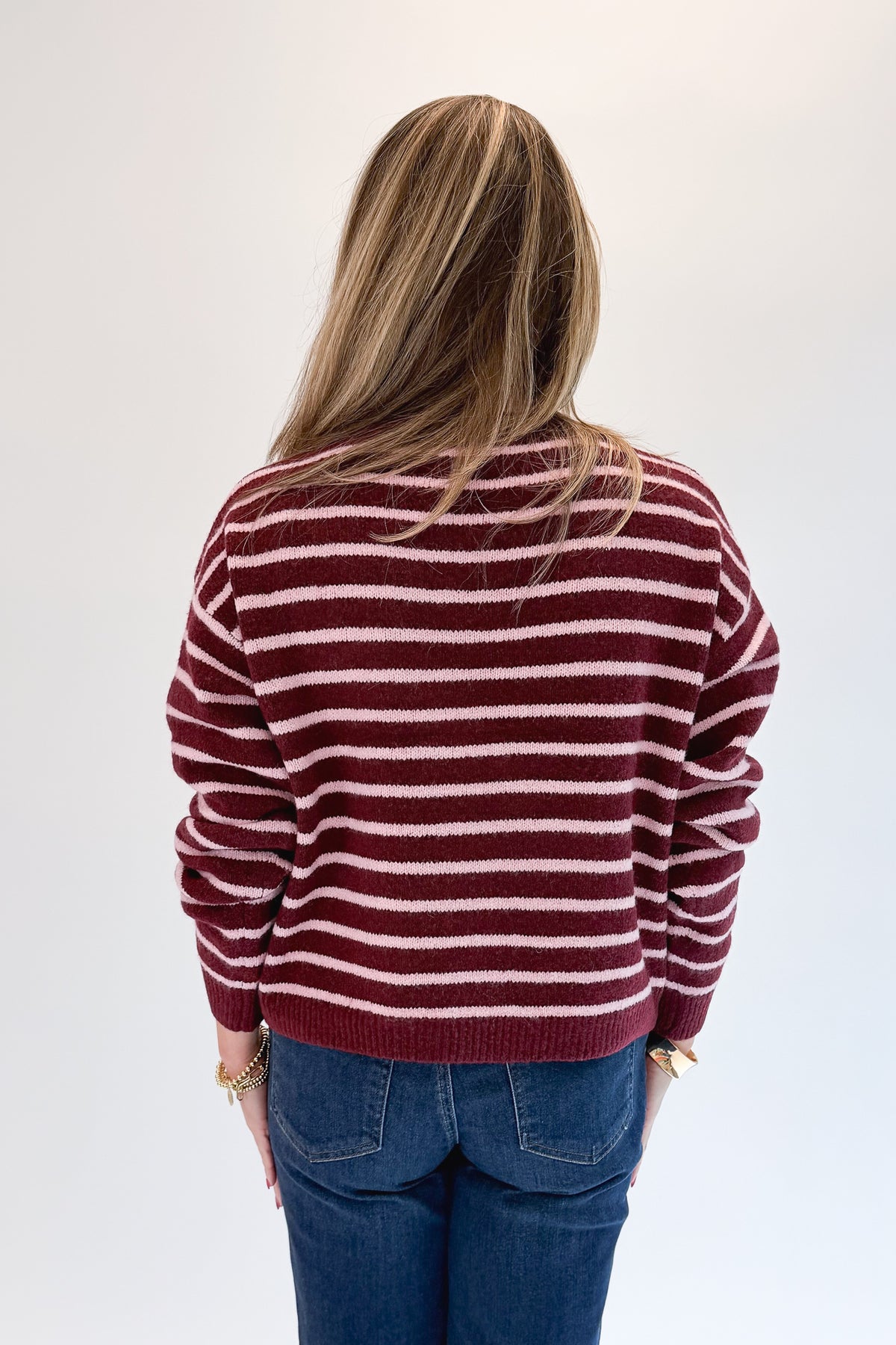 Back view of woman wearing Swagger Boutique’s Burgundy &amp; Pink Striped Sweater with jeans against white studio background.