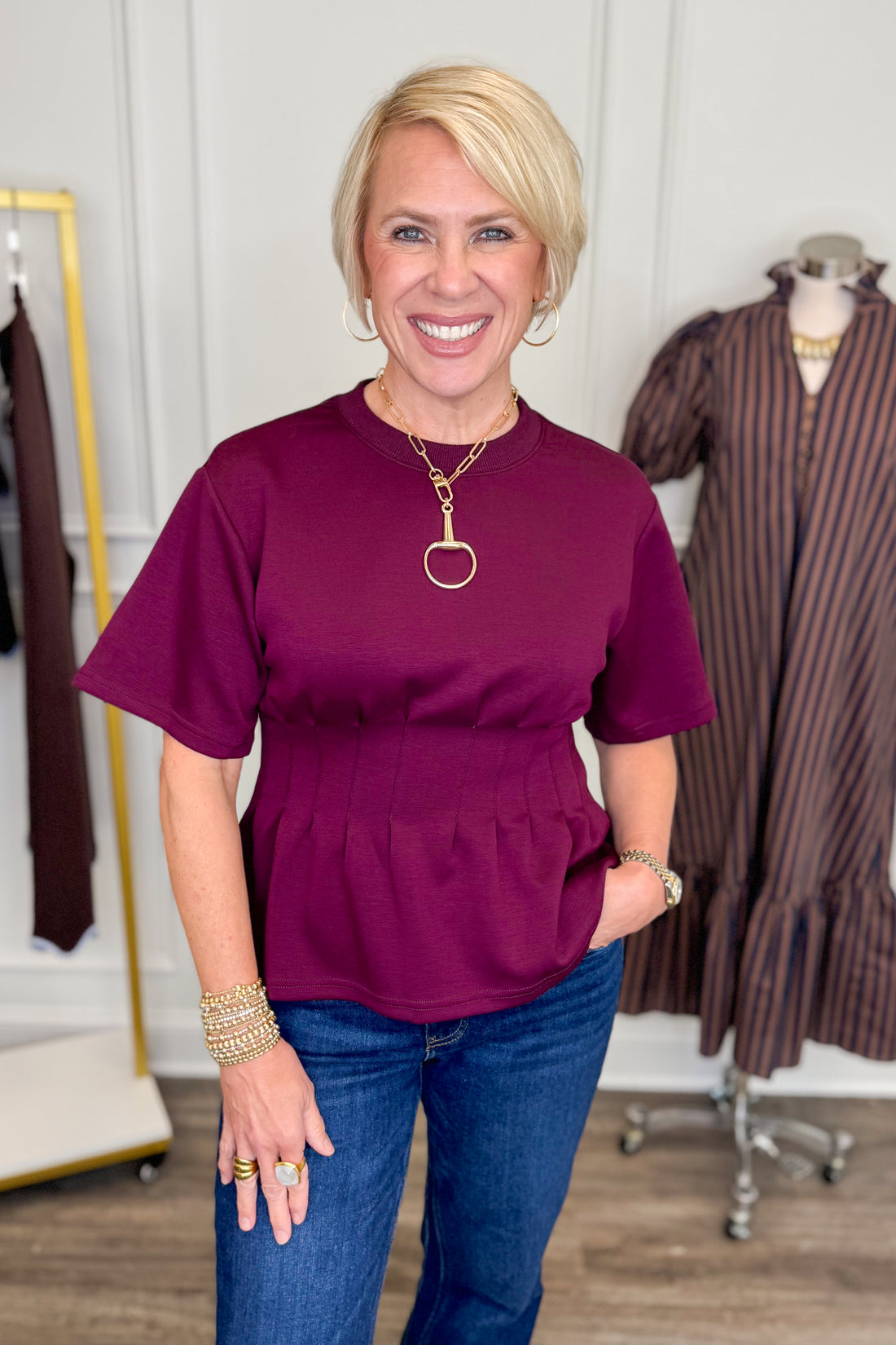 Front view of woman wearing Swagger Boutique’s Burgundy Pleat Waist Short Sleeve Top by Entro with jeans with clothing hanging in the background.