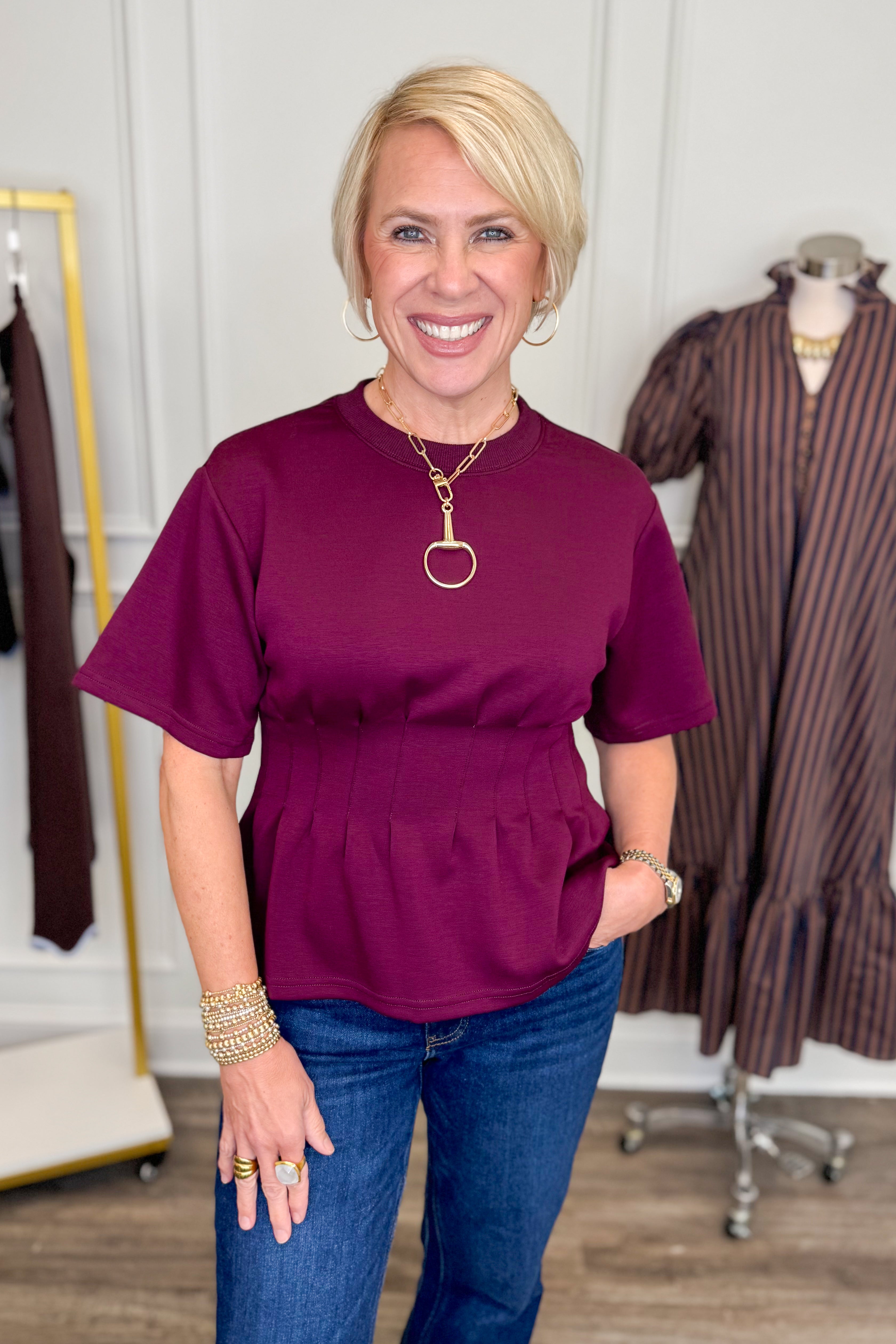 Front view of woman wearing Swagger Boutique’s Burgundy Pleat Waist Short Sleeve Top by Entro with jeans with clothing hanging in the background.