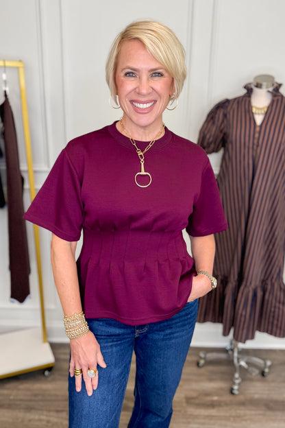 Front view of woman wearing Swagger Boutique’s Burgundy Pleat Waist Short Sleeve Top by Entro with jeans with clothing hanging in the background.