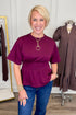 Front view of woman wearing Swagger Boutique’s Burgundy Pleat Waist Short Sleeve Top by Entro with jeans with clothing hanging in the background.