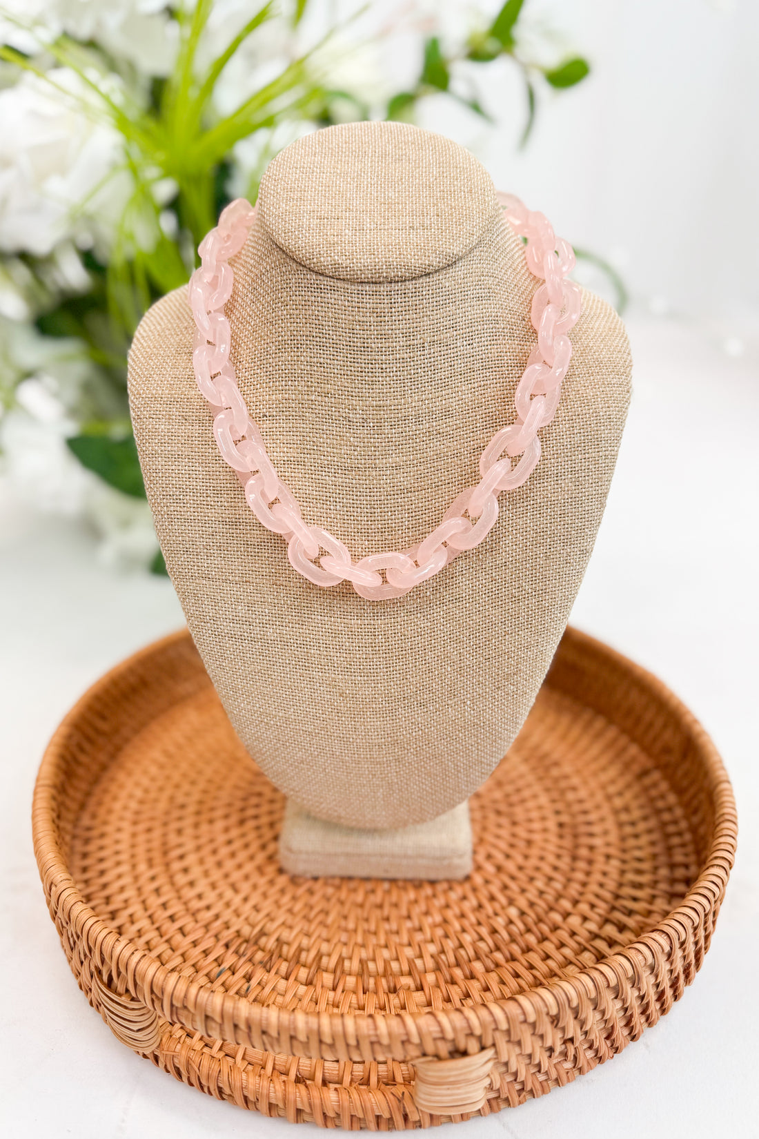 Pink necklace on a beige jewelry display with a woven basket and blurred flowers in the background