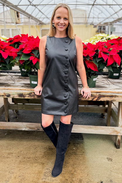 Front view of woman wearing Black Faux Leather Scallop Detail Dress with tall boots against around holiday decor.