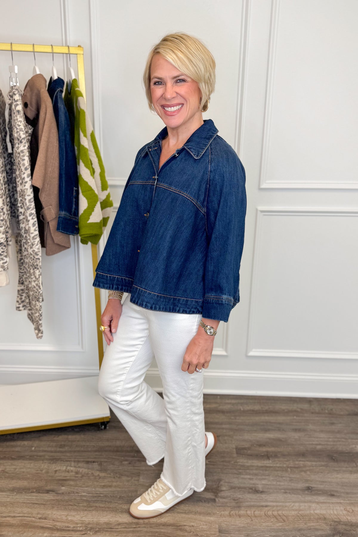 Side view of woman wearing Swagger Boutique’s Dark Denim Cape Top with jeans with clothing hanging in the background.