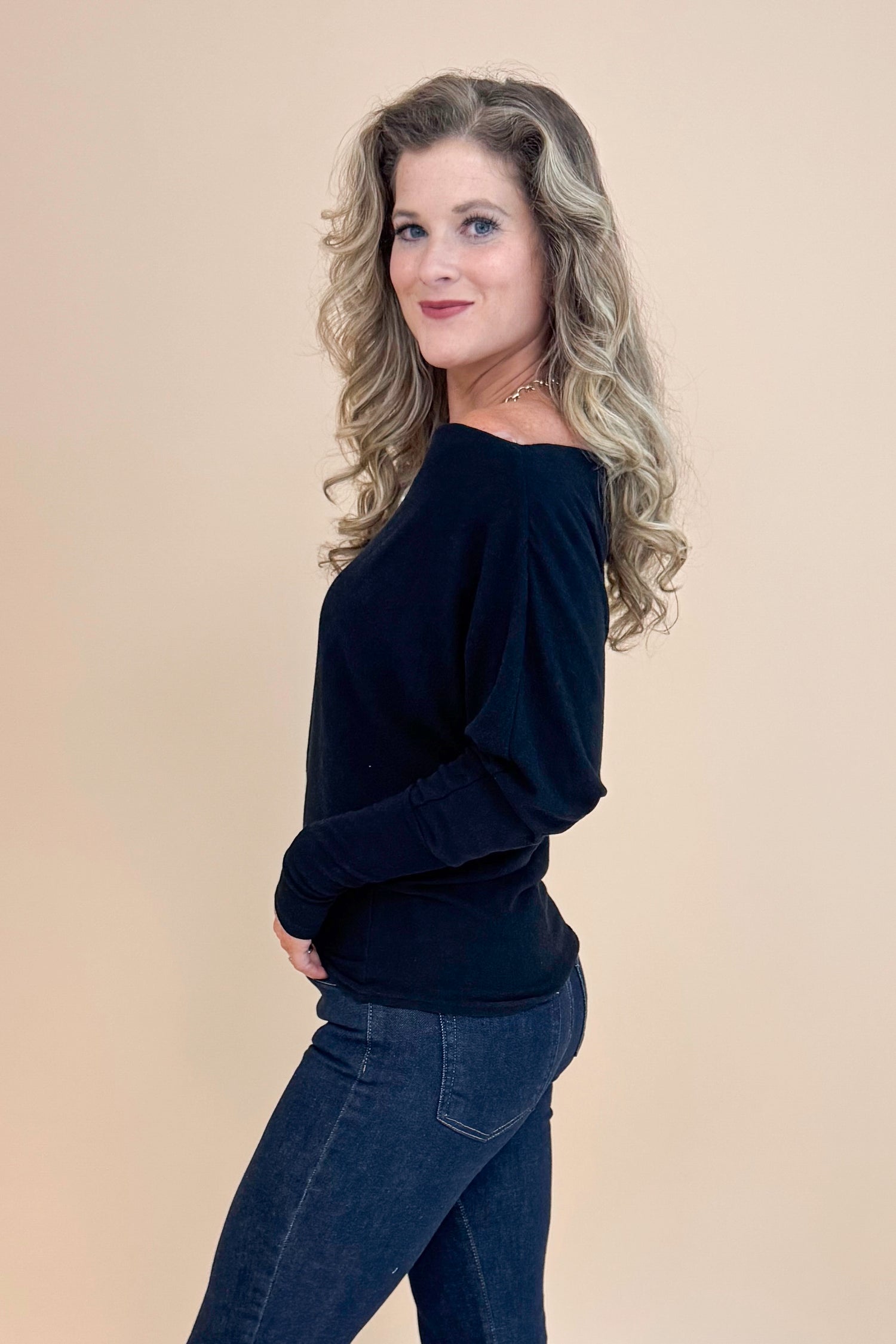 Side view picture of woman wearing Black Wide Neck Long Sleeve Top and jeans against a tan background.