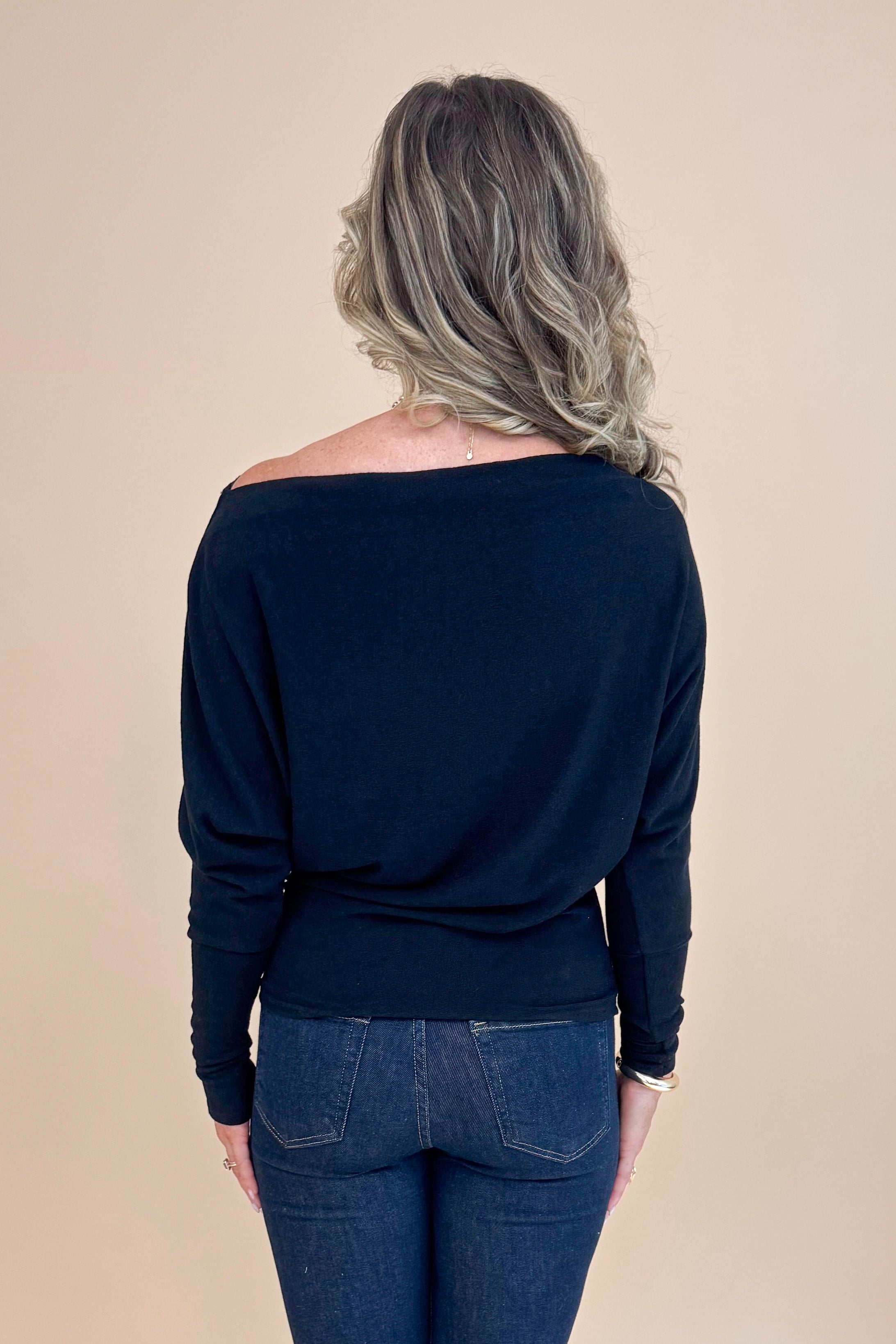 Back view picture of woman wearing Black Wide Neck Long Sleeve Top and jeans against a tan background.